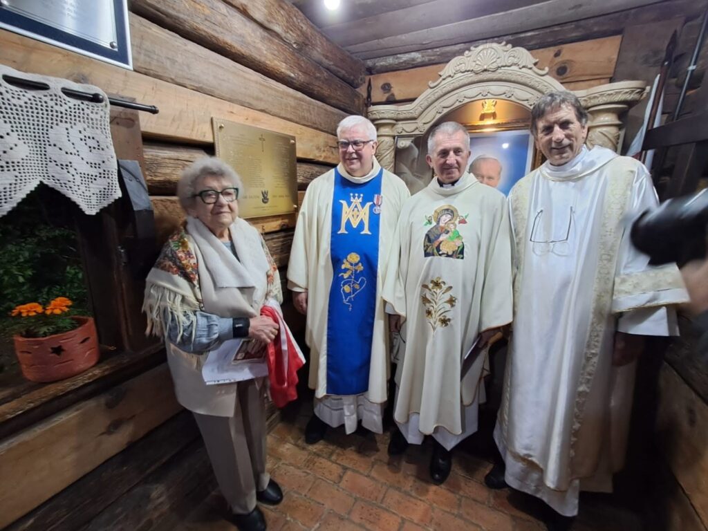 Kapłani w szatach liturgicznych i uczestniczka uroczystości w ludowej chuście w kaplicy Matki Bożej Częstochowskiej w Parku św. Jana Pawła II w Kurytybie; na ścianie nowa tablica