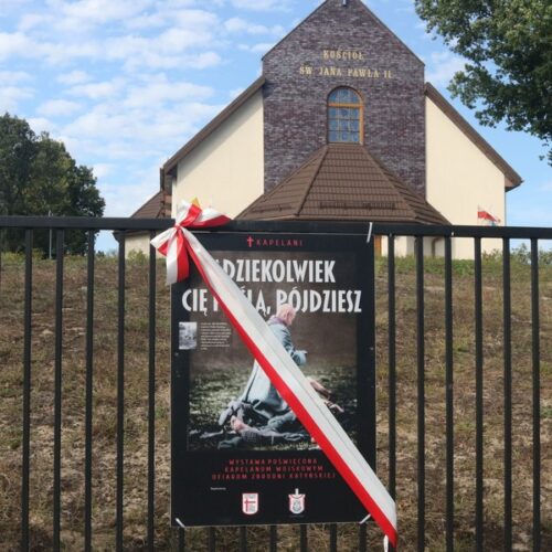 Plansza wystawy „Gdziekolwiek Cię poślą, pójdziesz” z biało-czerwoną wstęgą na ogrodzeniu kościoła pw. św. Jana Pawła II w Warszawie-Wesołej