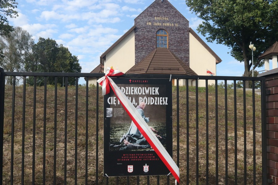 Plansza wystawy „Gdziekolwiek Cię poślą, pójdziesz” z biało-czerwoną wstęgą na ogrodzeniu kościoła pw. św. Jana Pawła II w Warszawie-Wesołej
